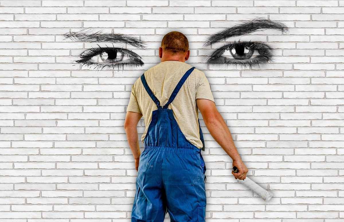 Homme en overalls peint un mur avec un rouleau. Œils dessinés sur le mur
