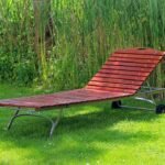 Chaise longue en bois rouge posée sur gazon, herbes hautes derrière