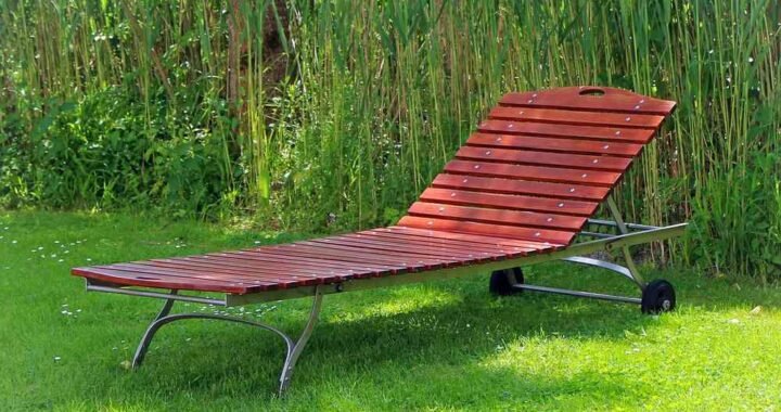 Chaise longue en bois rouge posée sur gazon, herbes hautes derrière