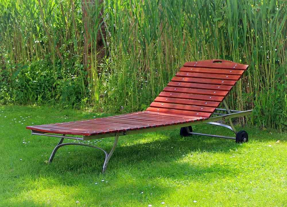 Chaise longue en bois rouge posée sur gazon, herbes hautes derrière